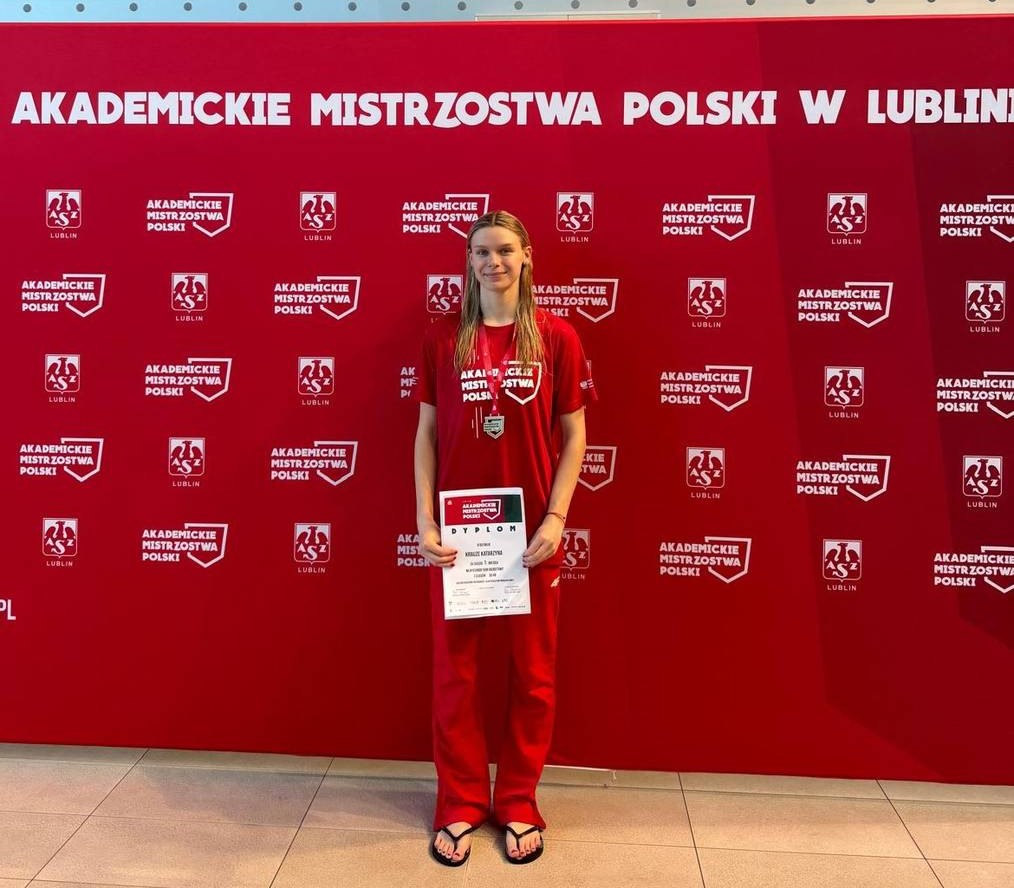 Kobieta trzymająca w rękach dyplom stoi na tle baneru Akademickich Mistrzostw Polski w Lublinie